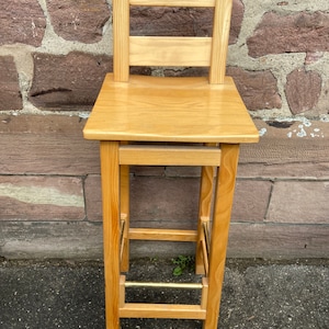 May include: A wooden bar stool with a light brown finish and a brass footrest. The stool has a simple design with a rectangular seat and a back with two horizontal slats.
