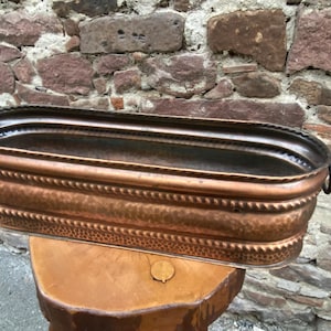 May include: An antique copper planter with a rectangular shape and decorative embossed detailing. The planter has a dark patina and is supported by a wooden stand. The copper planter is ideal for displaying plants or flowers.