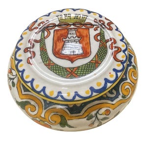 May include: A round, ceramic trinket box with a detailed, hand-painted design. The lid features a crest with a castle and a decorative border. The sides have a floral and scroll pattern in yellow, green, and blue. The box is white.