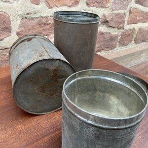 May include: Three antique metal containers with a weathered appearance. The containers are cylindrical, with one lying on its side, one upright, and one open. The metal has a dull, grey colour with signs of rust, suggesting age and use. The background is a brick wall.