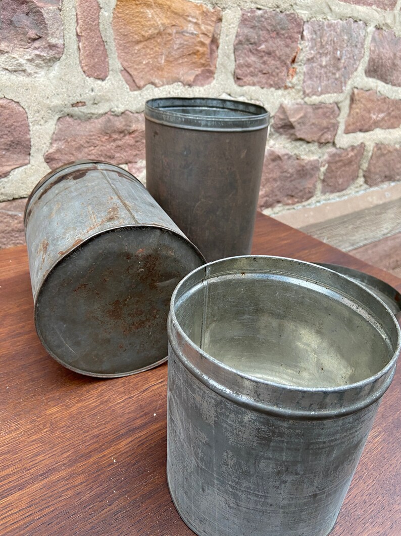 May include: Three antique metal containers with a weathered appearance. The containers are cylindrical, with one lying on its side, one upright, and one open. The metal has a dull, grey colour with signs of rust, suggesting age and use. The background is a brick wall.