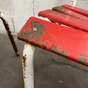 May include: Close-up of a red metal chair with white metal legs. The chair has a slatted seat and shows signs of rust and wear.