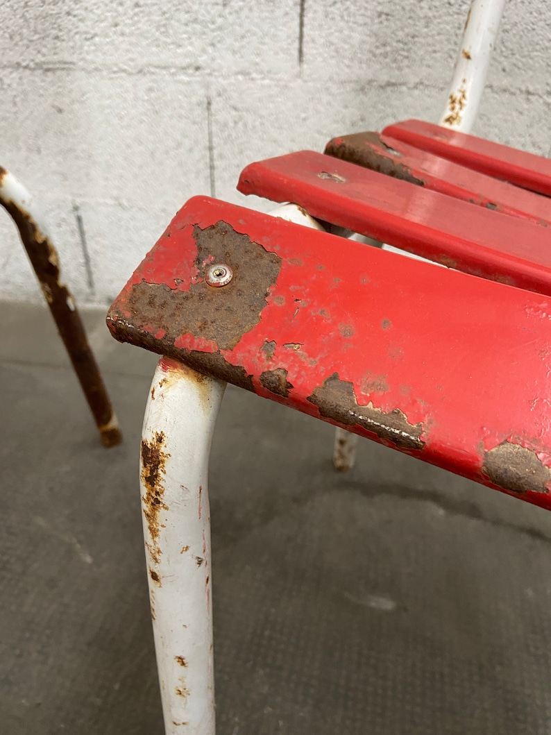 May include: Close-up of a red metal chair with white metal legs. The chair has a slatted seat and shows signs of rust and wear.