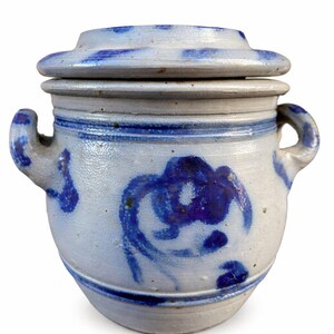 May include: A ceramic jar with a lid, featuring a white base and blue painted floral designs. The jar has two handles and a blue stripe around the middle. The lid also has blue accents.