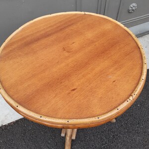 Puede incluir: Una mesa redonda de madera con un acabado de madera natural y un borde de bamb&uacute; tejido. La mesa tiene una base de tr&iacute;pode hecha de bamb&uacute; tejido.