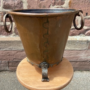 May include: A copper planter with three iron legs and two iron handles. The planter has a hammered texture and a dark patina.