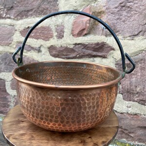 Könnte beinhalten: Ein gehämmerter Kupfertopf mit einem schwarzen Metallgriff. Der Topf hat eine strukturierte Oberfläche und steht auf einer Holzoberfläche.