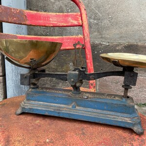 Peut inclure: Une balance vintage en métal bleu et noir avec deux bols en laiton. La balance est posée sur une chaise en métal rouge.