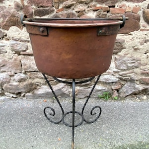 May include: A large, antique copper cauldron with a dark patina, supported by a wrought iron stand. The cauldron has a wide, open top with riveted handles. The stand features a decorative, scrolled base and three legs. The background is a weathered stone wall.