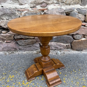 Op de afbeelding: Een ronde houten tafel met een sokkel en een gegraveerde, kruisvormige onderkant. De tafel is gemaakt van lichtbruin hout en heeft een gladde, gepolijste afwerking.