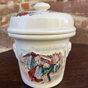Peut inclure: Pot en céramique crème avec couvercle, orné d'une scène peinte de danseurs en costumes traditionnels. Le couvercle est décoré de motifs floraux rouges, bleus et verts. Le pot a deux petites poignées.