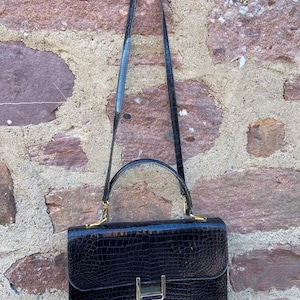 Peut inclure: Sac à main rectangulaire noir avec un motif texturé en forme de crocodile. Le sac est doté d'une poignée supérieure et d'une longue bandoulière. Des éléments dorés ornent la fermeture et les attaches de la bandoulière. Le sac est suspendu contre un mur en pierre rugueuse.