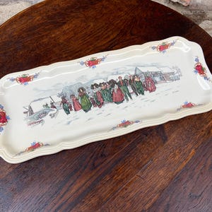 Puede incluir: Bandeja rectangular crema con borde festoneado. La bandeja presenta una escena invernal con figuras con trajes de época y edificios. Diseños florales y foliares adornan las esquinas y los bordes.