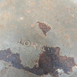 May include: Close-up of a weathered, circular metal object with the name "LORI" stamped on it. The surface is a muted grey with patches of rust and discoloration, suggesting age and exposure to the elements.