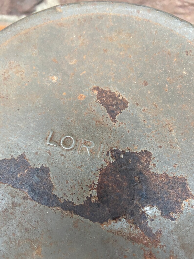 May include: Close-up of a weathered, circular metal object with the name "LORI" stamped on it. The surface is a muted grey with patches of rust and discoloration, suggesting age and exposure to the elements.