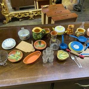 Könnte beinhalten: Eine Sammlung von Miniatur-Küchenutensilien, darunter ein blau-weißer Teller, ein Holzbrett, ein Metallsieb, eine rot-orangefarbene Plastikschüssel, eine blaue Plastikkelle, ein blaues Plastiksieb, ein blauer Plastiklöffel, ein rot-weißes Holznudelholz, eine silberne Gabel und ein Löffel sowie verschiedene Keramik-Schalen und Krüge.