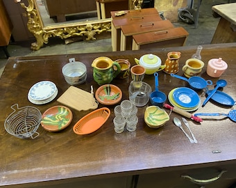 May include: A collection of miniature kitchenware, including a blue and white plate, a wooden cutting board, a metal colander, a red and orange plastic bowl, a blue plastic ladle, a blue plastic strainer, a blue plastic spoon, a red and white wooden rolling pin, a silver fork and spoon, and a variety of ceramic bowls and pitchers.