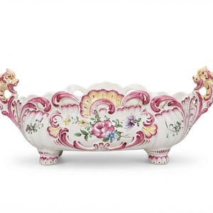 Pode incluir: Um centro de mesa de cerâmica alongado com base branca e volutas pintadas à mão em rosa e amarelo. O recipiente possui duas alças ornamentadas em forma de criaturas míticas e apoia-se sobre quatro pés pequenos com motivos florais.