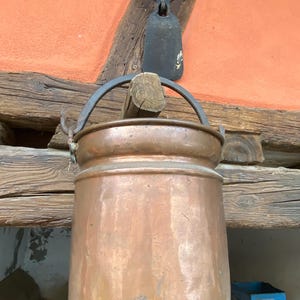 May include: A copper-colored metal pot with a rounded body and a curved handle. The pot is suspended from a wooden beam, with other rustic tools and architectural elements visible in the background. The pot has a vintage, antique appearance.