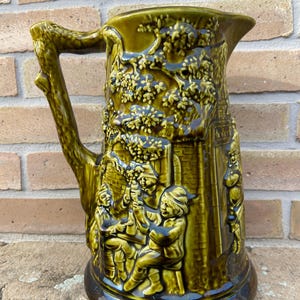 Peut inclure: Un pichet en céramique vert avec un motif d'arbre et de personnes. Le pichet a une poignée brune en forme de branche d'arbre.