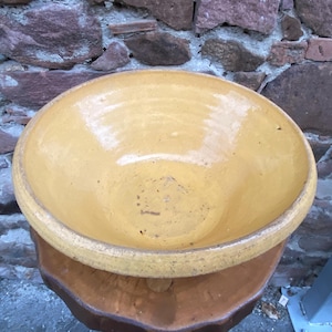 May include: A large, yellow ceramic bowl with a wide rim. The bowl is set on top of a smaller, brown ceramic bowl. The background is a stone wall. The bowl is likely used for serving food or as a decorative piece.