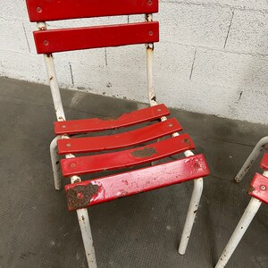 May include: Two red metal chairs with white painted legs. The chairs have a slatted seat and back. The paint is chipped and shows signs of rust.