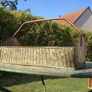 Puede incluir: Un macetero vintage de metal galvanizado con un asa de cobre. El macetero tiene un diseño corrugado y está desgastado con manchas de óxido.