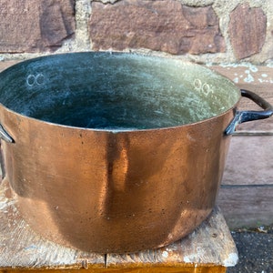 Può includere: Una grande pentola di rame con due manici neri. La pentola ha un aspetto usurato e invecchiato e si trova su una superficie di legno.