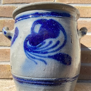 Op de afbeelding: Een grote, grijze steengoed pot met twee handvatten. De pot is versierd met een blauw en wit bloemenmotief.