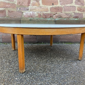 Puede incluir: Una mesa de centro redonda con una tapa blanca y un marco de madera y cuatro patas. La mesa está hecha de madera y tiene un acabado de madera clara.