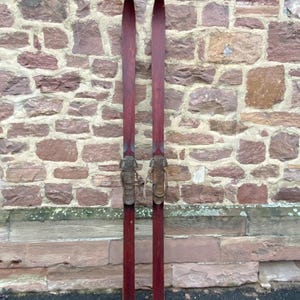 May include: A pair of vintage wooden skis with metal bindings. The skis are a deep reddish-brown color and stand upright against a weathered brick wall. The tips are pointed, and the bindings are a light brown color.