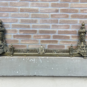 May include: Ornate brass fireplace fender with two decorative finials. The fender has a detailed design with scrolls and floral motifs.
