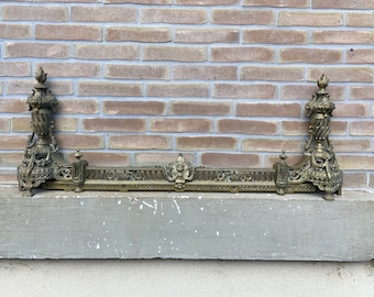 May include: Ornate brass fireplace fender with two decorative finials. The fender has a detailed design with scrolls and floral motifs.