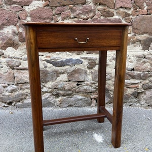 Könnte beinhalten: Ein dunkelbrauner Holz-Beistelltisch mit einer Schublade und einem Messinggriff. Der Tisch hat ein schlichtes Design mit geraden Beinen und einer rechteckigen Tischplatte.