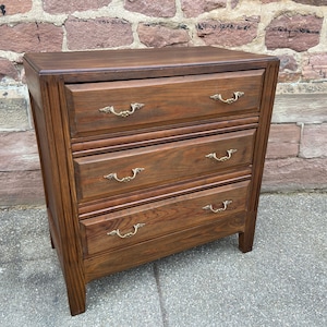 Op de afbeelding: Een donkerbruine houten commode met drie laden en messing handgrepen. De commode heeft een eenvoudig ontwerp en is gemaakt van massief hout.