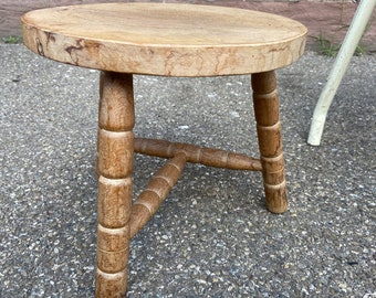 May include: A round wooden stool with three turned legs. The legs have a decorative knob at the bottom. The stool is made of light brown wood and has a natural finish.