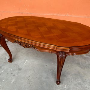 Op de afbeelding: Ovale salontafel van donkerbruin hout. Het tafelblad heeft een parketpatroon. De tafel heeft sierlijke, gebogen poten en decoratieve snijwerk. Een klassiek meubelstuk, ongeveer 150 cm lang.