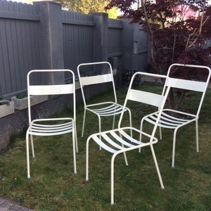 May include: A set of four off-white metal chairs featuring a tubular frame, a horizontal backrest slat, and a seat constructed from multiple thin, parallel metal bars. The chairs have four slender legs and a simple, functional design.