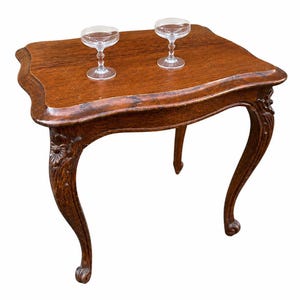 Puede incluir: Una mesa auxiliar de madera marrón con una superficie rectangular de bordes curvos y cuatro patas cabriolé con detalles florales tallados en la parte superior. Dos copas de cristal transparente de tallo corto están sobre la mesa.