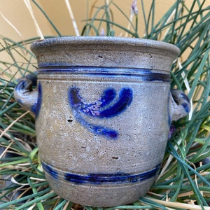 Op de afbeelding: Een grijze steengoed pot met blauwe en witte beschilderde ontwerpen. De pot heeft twee handvatten en een brede opening.