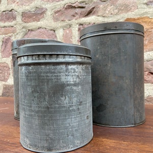 May include: Three antique metal storage tins with lids. The cylindrical tins have a distressed, weathered appearance in a dark grey colour. They are arranged on a wooden surface against a brick wall background.