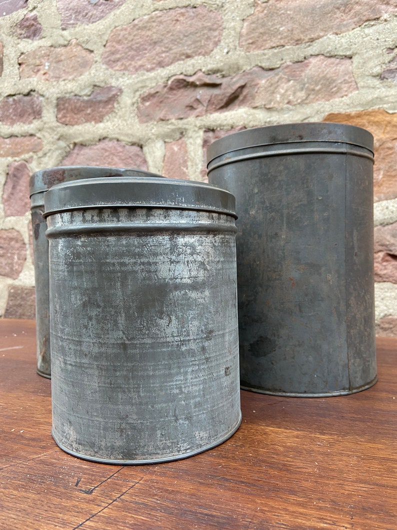 May include: Three antique metal storage tins with lids. The cylindrical tins have a distressed, weathered appearance in a dark grey colour. They are arranged on a wooden surface against a brick wall background.