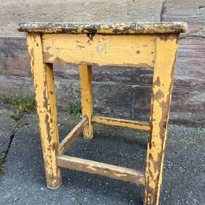 Puede incluir: Un pequeño taburete de madera amarillo con un acabado desgastado. El taburete tiene cuatro patas y una parte superior rectangular simple.
