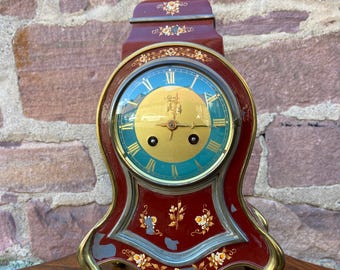 May include: A red and gold mantle clock with a painted floral design. The clock has a keyhole on the side and a key on the table below.