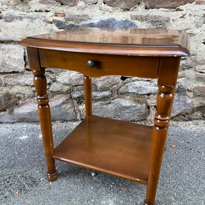 Puede incluir: Una mesa auxiliar de madera marrón oscuro con un cajón y un estante inferior. La mesa tiene patas torneadas y una parte superior redondeada.