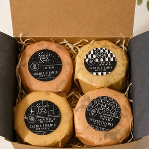 May include: A brown cardboard box containing four round shower steamers wrapped in paper. Each steamer has a black circular label with the text Soaps & Suds, the scent name, and Min Wt: 40g. Scents include Lavender, Tea Tree & Lemongrass, Grapefruit & May Chang, and Geranium & Rosemary.