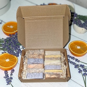 May include: A selection of handmade soaps presented in a cardboard box. The soaps are rectangular and feature diverse colours and textures. Sliced oranges and lavender sprigs surround the box, creating a natural and aromatic display.