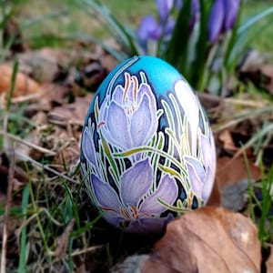 Peut inclure: Un œuf de Pâques décoré avec un motif floral de fleurs de crocus violettes sur un fond bleu et blanc. L'œuf repose sur un lit de feuilles brunes et d'herbe verte, avec d'autres fleurs violettes en arrière-plan.