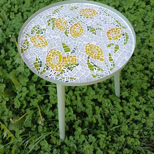 Puede incluir: Una pequeña mesa redonda de mosaico con marco y patas verde claro. La mesa presenta un diseño de mosaico de flores amarillas y hojas verdes sobre un fondo azul claro. La mesa está sobre trébol verde.