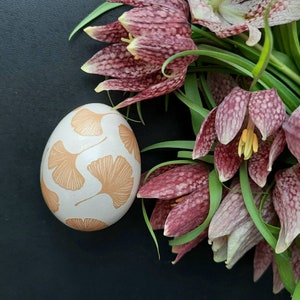 Op de afbeelding: Een glad, gebroken wit ei versierd met een herhalend patroon van lichtoranje ginkgobladeren, geplaatst naast een tros klokvormige kievitsbloemen met een paars en wit geblokt patroon en groene stengels.